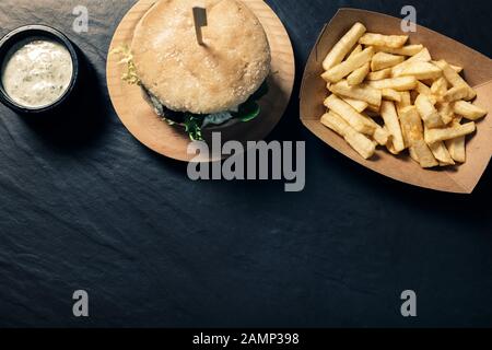 Gourmet-Burger mit Salat auf schieferem Hintergrund mit pommes frites in einem Pappkarton und einem Krug mit Soße, Foodie Lifestyle-Konzept Stockfoto