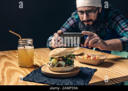 Koch fotografiert mit dem Telefon seines veganen Burgers mit Salat und Sauce, Pommes mit Ketchup und gesundem Getränk auf einem Holztisch, Technologie und Stockfoto