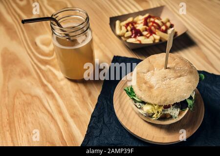 Veganer Gourmet-Burger mit Salat und Soße auf einem Holztisch mit pommes frites mit Ketchup in einem Pappkarton und einem gesunden Getränk, veggie Essen und Stockfoto