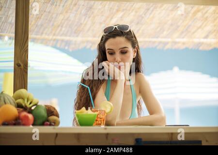 Traurige junge Frau, die allein an der Strandbar trinkt, lehnt sich an die Theke und denkt Stockfoto