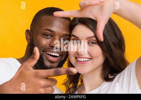 Positives Paar mit mehreren Rassen, das durch den Fingerrahmen blickt und über gelbem Hintergrund posiert Stockfoto