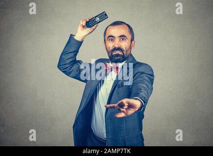 Halb Körperlänge Porträt eines Mannes, der eine tv-Fernbedienung wirft, die Sie auf Ihre Kamera schaut, erschrocken und wütend. Gebärdetes Modell mit gemischtem Rennen, isoliert auf grauem Rücken Stockfoto