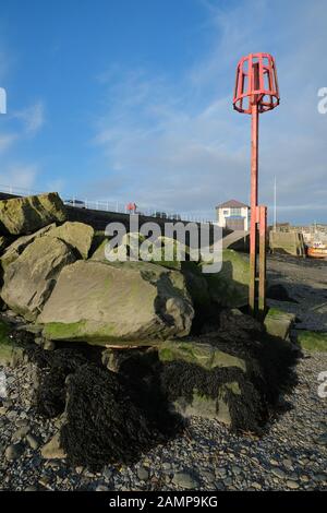 Aberystwyth Harbour: Roter Gezeitenmarker vor blauem Himmelshintergrund und großen Felsbrocken Stockfoto