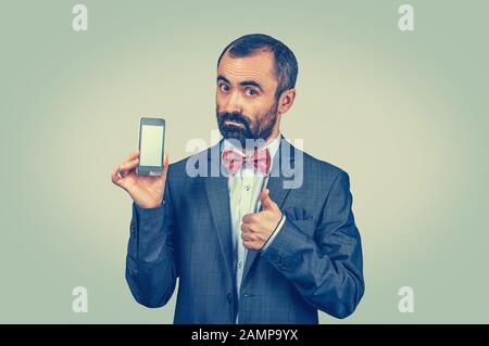 Geschäftsmann, der mit Daumen nach oben geht, wie Geste mit der Hand und halten, dass der Telefonbildschirm in der anderen Hand angezeigt wird. Gemischtes Rennbären-Modell isoliert auf Grüngelb Stockfoto