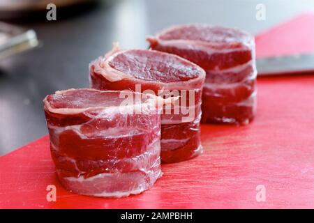 Rohes Filet von Hereford Irish Beef Filet Steak in parmaschinken auf einem roten Hackbrett mit Messer im Hintergrund eingewickelt. Stockfoto