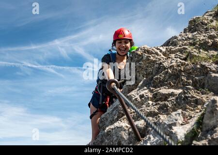 Lachende junge Frau mit Kletterhelm und auf dem mit Seil gesicherten Felsen, weibliche Kletterin auf einem Klettersteig, Mittenwald über Ferrata, Karwendel Stockfoto