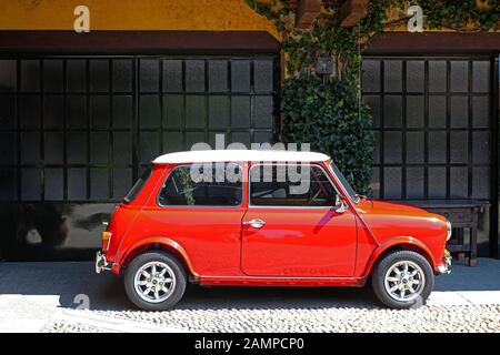 Red Austin Mini mit weißem Dach steht an einem sonnigen Tag auf einem gepflasterten Innenhof vor einer langen Garagentür. Stockfoto