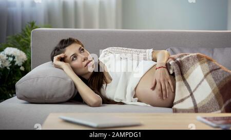 Besorgt über zukünftige Mama, die auf dem Sofa liegt, sanft den Bauch reibt und an die Zukunft denkt Stockfoto