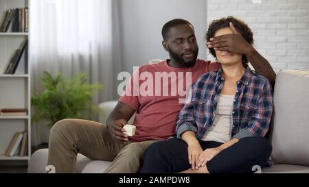 Afro-amerikanischer Mann, der die Augen vor dem Verlobten schließt, der überrascht und dabei Geschenk in der Hand hält Stockfoto