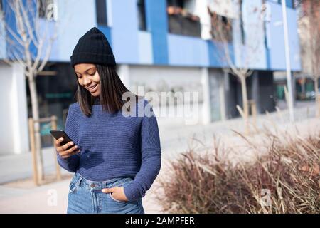 Porträt der jungen afroamerikanischen Frau, die eine Wollkappe trägt, die auf der Straße steht, während sie ein Mobiltelefon verwendet Stockfoto