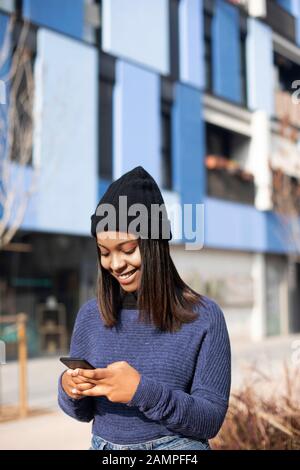 Porträt der jungen afroamerikanischen Frau, die eine Wollkappe trägt, die auf der Straße steht, während sie ein Mobiltelefon verwendet Stockfoto