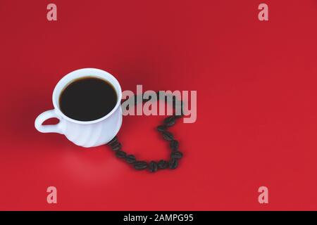 Weiße Tasse voller Kaffee und eine Herzform mit Kaffeebohnen auf hellrotem Hintergrund. Liebe Kaffeekonzept Stockfoto