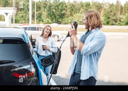 Transport. Junge Paare, die mit dem Auto unterwegs sind, haben einen Stopp an der Ladestation. Freund spielt mit einem Stecker, der auf die Freundin schaut, die heißen Kaffee trinkt Stockfoto