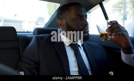 Nachdenklicher afroamerikanischer Mann, der mit Alkoholgetränk auf dem Rücksitz des Autos sitzt Stockfoto