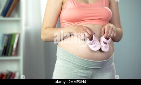 Schwangere Frau erwartet kleines Mädchen, hält rosafarbene Schuhe in der Nähe des Bauches, Babydusche Stockfoto