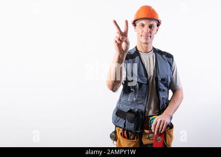 Erbauer zeigt ein Ziegenschild. In Arbeitskleidung und Harthut. Auf hellgrauem Hintergrund. Stockfoto