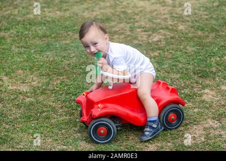 Kleiner Junge, der Kamm hält, während er auf einem roten Spielzeugauto im Garten sitzt. Stockfoto