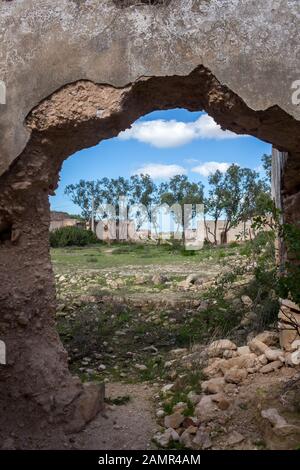 Mauer eines verlassenen Gebäudes (Kasbah) mit einem großen Loch und Blick auf eine Herbstwiese mit einer Baumreihe im Hintergrund. Strahlend blauer Himmel, weiß cl Stockfoto