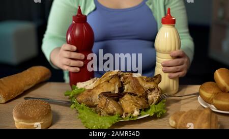 Fette Dame, die Flaschen Ketchup und Mayonnaise hält, bereit zum Verzehr von Braten Huhn Stockfoto