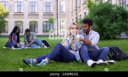 Junge Paare sitzen auf dem Rasen auf dem Campus und sehen Bilder auf dem Smartphone Stockfoto