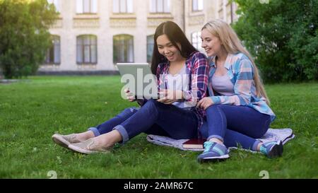 Zwei Studentinnen sitzen auf Gras, lachen auf Bildern in Laptop, einkaufen Stockfoto