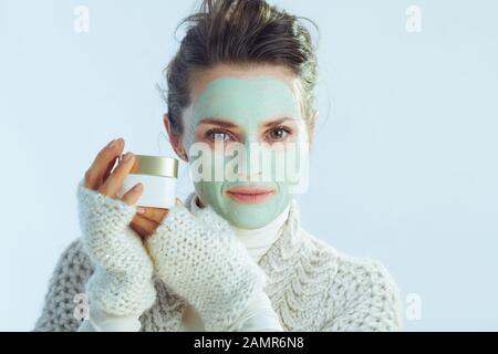 Moderne Frau im mittleren Alter mit Rollkragen-Pullover und Strickjacke mit grüner Gesichtsmaske, die einen kosmetischen Mixbecher auf winterhellblauem Hintergrund zeigt. Stockfoto