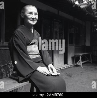 1960, historische, alte japanische Dame draußen sitzen auf einer Bank das Tragen eines Kimonos, die traditionelle Kleidung von Japan. In der Regel aus Seide, sie große ärmel, gehen Sie von den Schultern bis zu den Fersen und wird von einem OBI-Gehalten, einen breiten Gürtel. Stockfoto