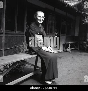 1960, historische, alte japanische Dame außerhalb einer bambo gebaute Hütte auf einer Bank das Tragen eines Kimonos, die traditionelle Kleidung der Japan sitzen. In der Regel aus Seide, sie große ärmel, gehen Sie von den Schultern bis zu den Fersen und wird von einem OBI-Gehalten, einen breiten Gürtel. Stockfoto