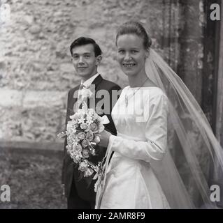 1960er Jahre, historisches, junges Hochzeitspaar für Erwachsene, lächelnde Frau in ihrem Hochzeitskleid, mit Schleier, der einen Blumenstrauß außerhalb einer Kirche, England, Großbritannien, hält. Stockfoto