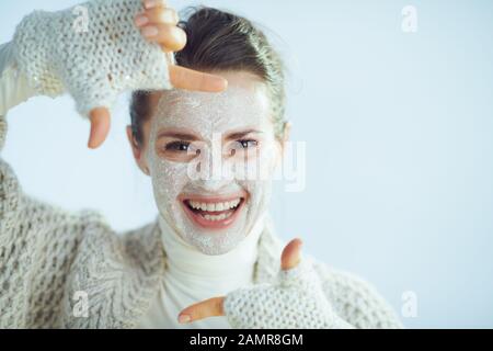 Lächelnde, elegante Frau mit Rollkragen-Pullover und Strickjacke mit weißer Gesichtsmaske, die mit den Händen auf hellblauem Winterhintergrund umrahmt wird. Stockfoto