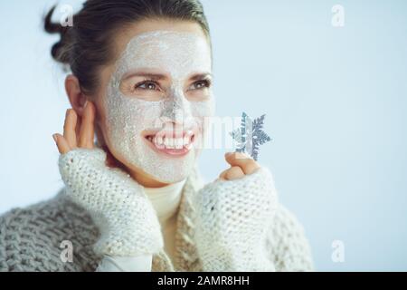 Lächelnde, stilvolle Frau im mittleren Alter mit Rollkragen-Pullover und Strickjacke mit weißer Gesichtsmaske als Teil der Winter-Hautpflege, die Schneeflocke mit Blick auf den Cop hält Stockfoto