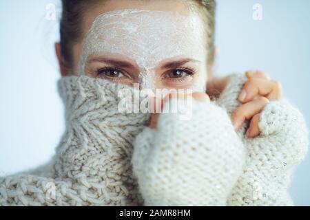 Elegante Frau in Rollkragen-Pullover und Strickjacke mit weißer Gesichtsmaske, die sich hinter der Kleidung auf hellblauem Winterhintergrund versteckt. Stockfoto