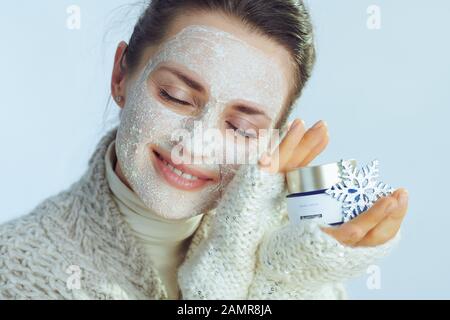 Fröhliche elegante Frau im Rollkragen-Pullover und Strickjacke mit weißer Gesichtsmaske, die auf hellblauem Hintergrund einen kosmetischen Mixbecher mit Schneeflocke zeigt. Stockfoto