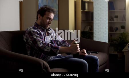 Bärtiger Mann versucht, Bierflasche mit Gabel zu öffnen und sitzt auf der Couch im dunklen Raum Stockfoto