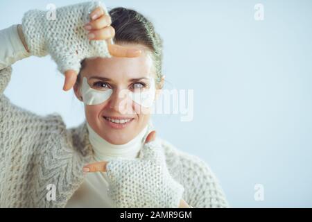 Porträt der fröhlichen, stylischen 40 Jahre alten Hausfrau mit Rollkragen-Pullover und Strickjacke mit kosmetischen Augenmustern, die mit den Händen am Winter hellblau umrahmt werden Stockfoto