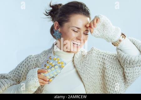 Fröhliche, stylische 40 Jahre alte Frau mit Rollkragen-Pullover und Strickjacke, die Blisterpackungen mit Pillen auf hellblauem Winterhintergrund hält. Stockfoto