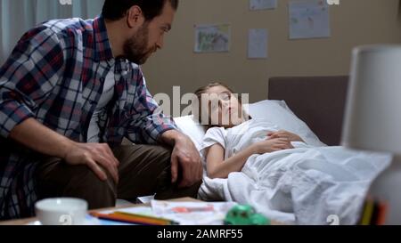 Vater spricht mit einer kleinen kranken Tochter, die im Bett liegt und sich um ein krankes Kind kümmert Stockfoto