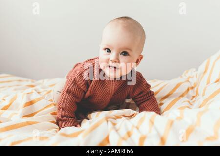 Schönes kleines Mädchen auf dem Bett in einem gemütlichen braunen Pullover lächelt. Konzept der Mutterschaft und Kindheit. Liebenswürdig sechs Monate altes Mädchen, das auf dem schlechten liegt und in die Kamera schaut. Stockfoto