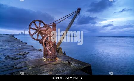 Alter rostiger manueller Dockkran am langen Pier, Der Great South Wall, mit verschwommenem Frachtschiff an den Docks in Dublin, Irland. Langzeitfotografie Stockfoto