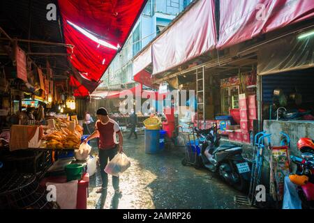 Khlong Toei Markt in Bangkok, Thailand mit Lebensmittelständen unter rotem Dach mit einem Mann, der Plastiktüten hält und bei einem Händler in einer Sonnenlicht-Straße kauft Stockfoto