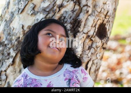 Ein 9 Jahre altes Mädchen träumt, während es sich an einen Baumstamm lehnt. Stockfoto