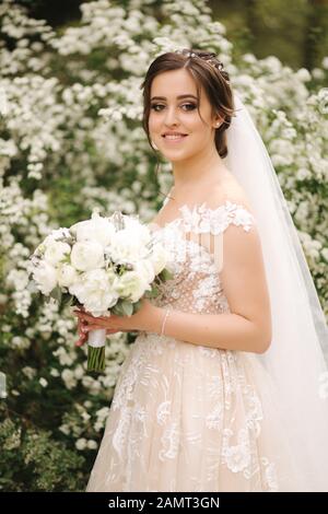 Bezaubernde Braut in Gorgeous Wedding Dress halten Bouquet und stehen vor der blühenden Bäume Stockfoto