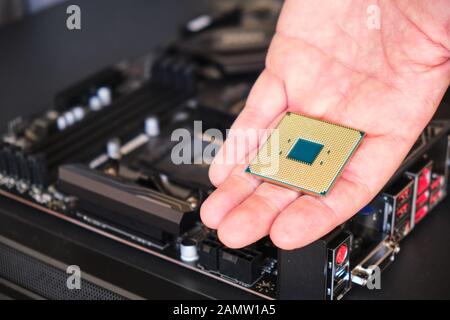 CPU (Central Processing Unit) in der Hand eines Mannes gehalten - Nahaufnahme, mit einer verschwommenen Hauptplatine im Hintergrund. Kopierbereich. Stockfoto