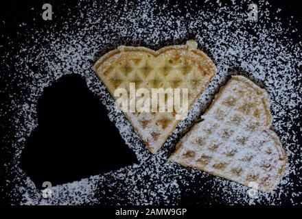 Zwei hausgemachte Waffeln in Form von Herzen liegen nebeneinander auf einem dunklen Teller und sind mit Zucker bestreut. Daneben fehlt eine Waffel Stockfoto