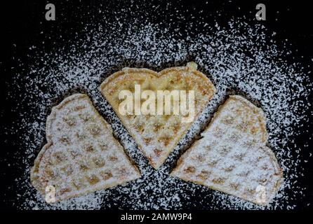 Drei hausgemachte Waffeln in Form von Herzen liegen nebeneinander auf einem dunklen Teller und sind mit Zucker bestreut Stockfoto