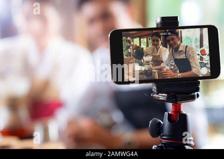 VLOG von jungen erwachsenen Asiatischen Eigentümer Unternehmer bewerten cafe Coffee Shop und Leben in sozialen Medien für Online Marketing mit Smart Phone im Cafe. Mit fo Stockfoto