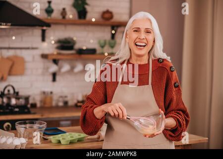 Grauhaarige lächelnde Dame, die in einer Schüssel leuchtende, peitschende Milch lächelt Stockfoto