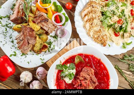 Verschiedene Gerichte auf Holztisch, Ansicht von oben. Gemüse Rüben Suppe, Lammrücken mit gebratenen Kartoffeln, gegrilltem Hühnchen Caesar Salat. Stockfoto