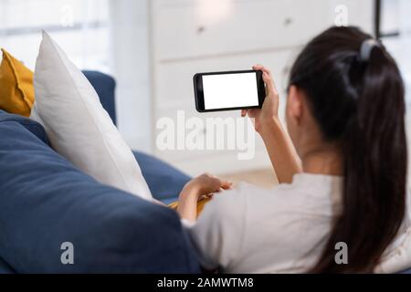 Junge asiatische Frau Holding mit leeren weißen Bildschirm smartphone, Liegen gemütlich auf dem Sofa im Wohnzimmer, Technologie elektronische Gadget Stockfoto