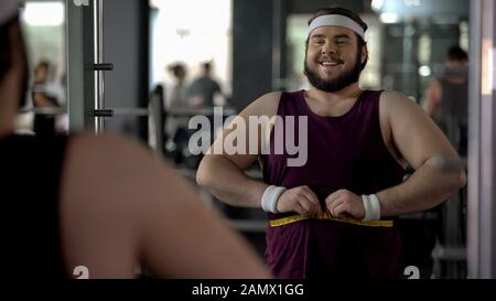 Glücklicher adipöser Mann, der die Taille misst, zufrieden mit den Ergebnissen des Trainingsprogramms Stockfoto